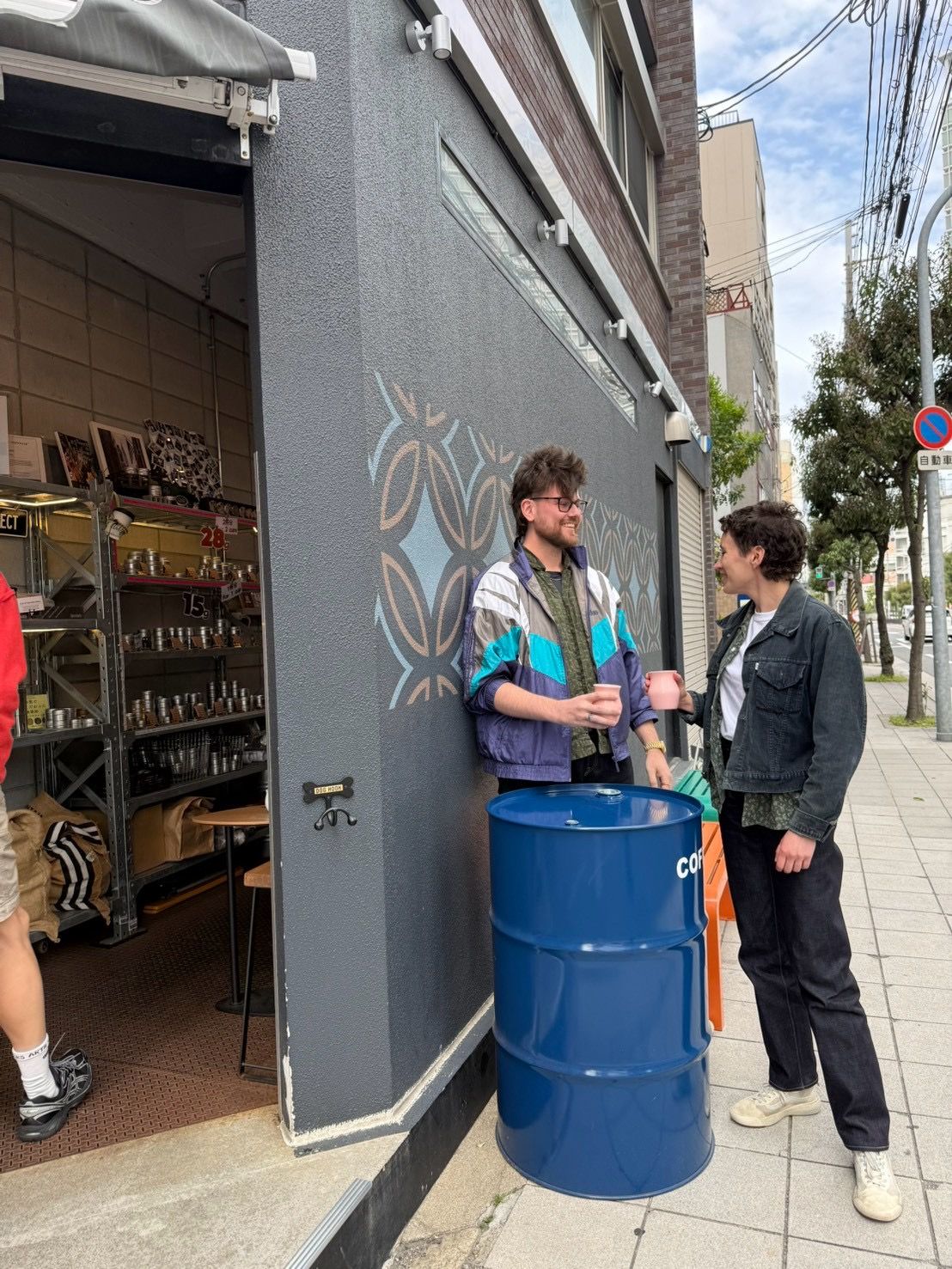 店先の青いドラム缶を囲み、カップを手に立ち話をする二人